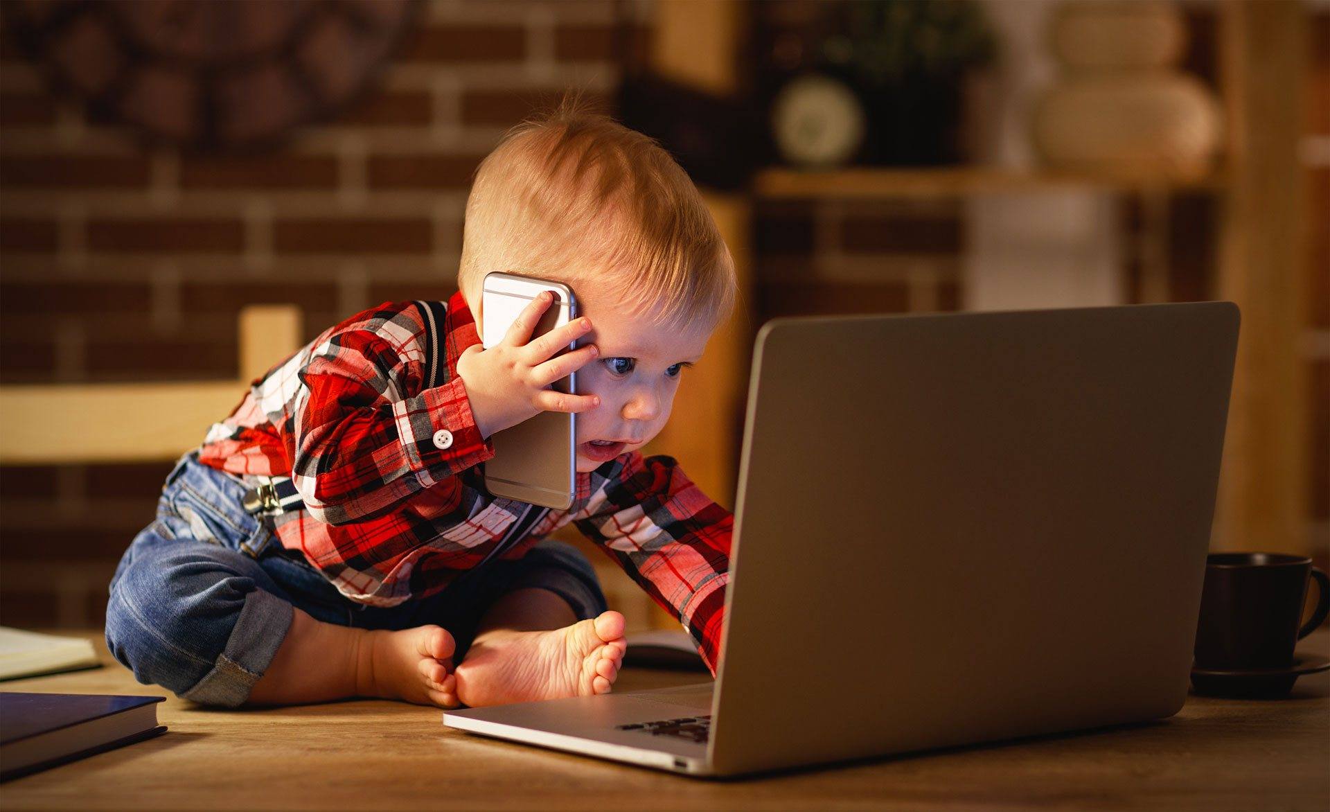 A baby sitting on the floor, holding a mobile phone while focused on a laptop