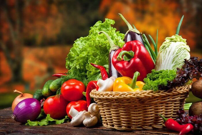 A basket overflowing with various fruits and vegetables sits on a wooden table